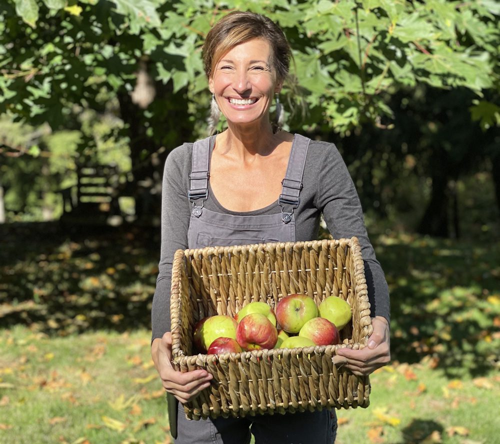 Nicole Apelian wearing Dovetail Workwear Freshly Overalls holding a basket of apples Nicole Apelian wearing Dovetail Workwear Freshly Overalls holding a basket of apples